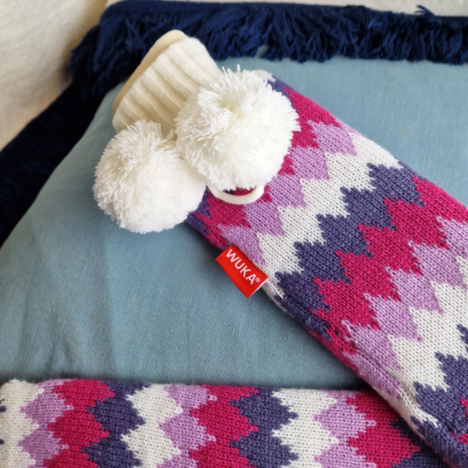 WUKA extra long hot water bottle resting on a cushion, showcasing vibrant pink and purple knitted cover with white pom poms.